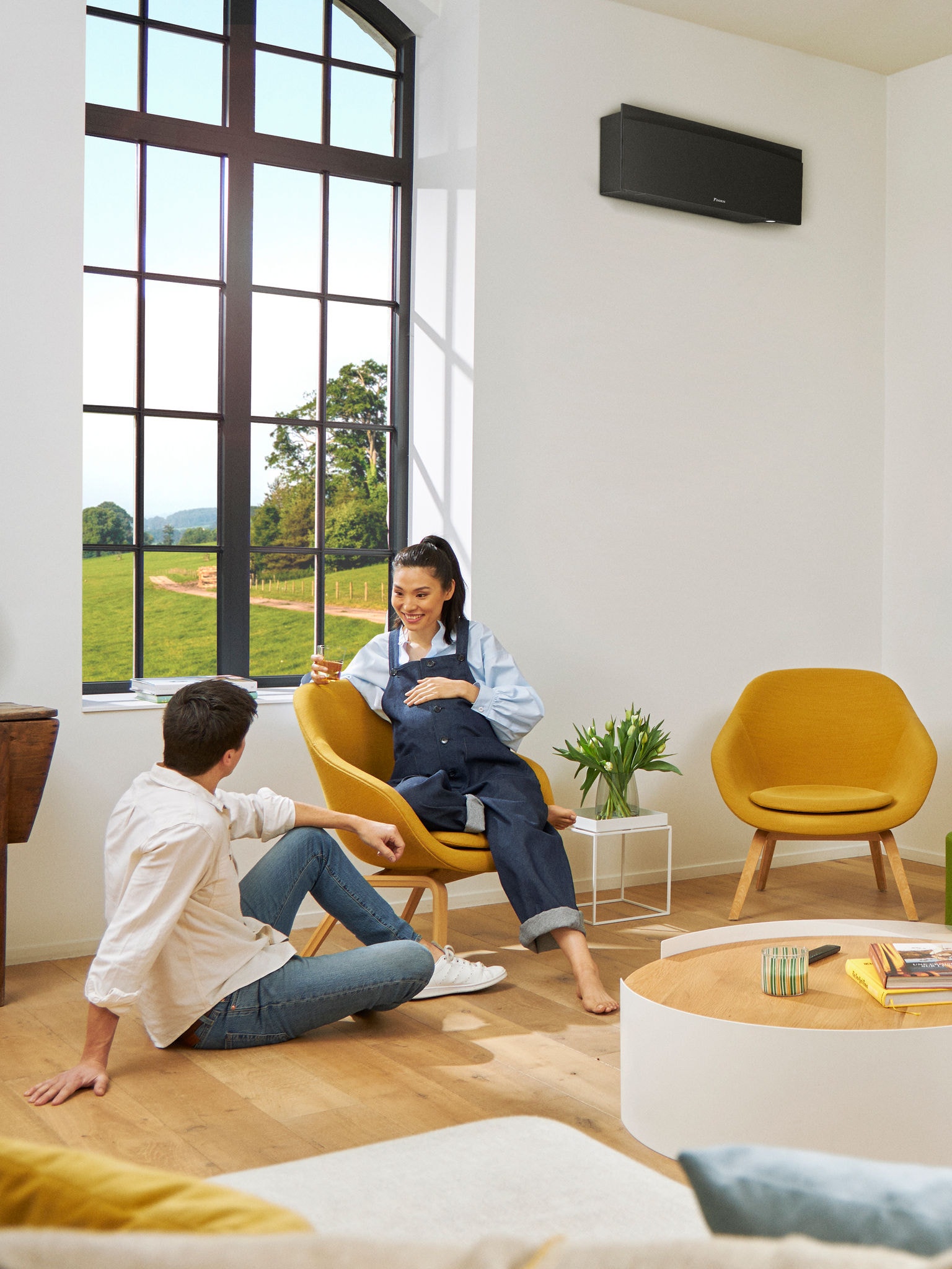 Een man en een vrouw zitten voor een groot raam in een lichte kamer met een zwarte wandwarmtepomp aan de muur achter hen