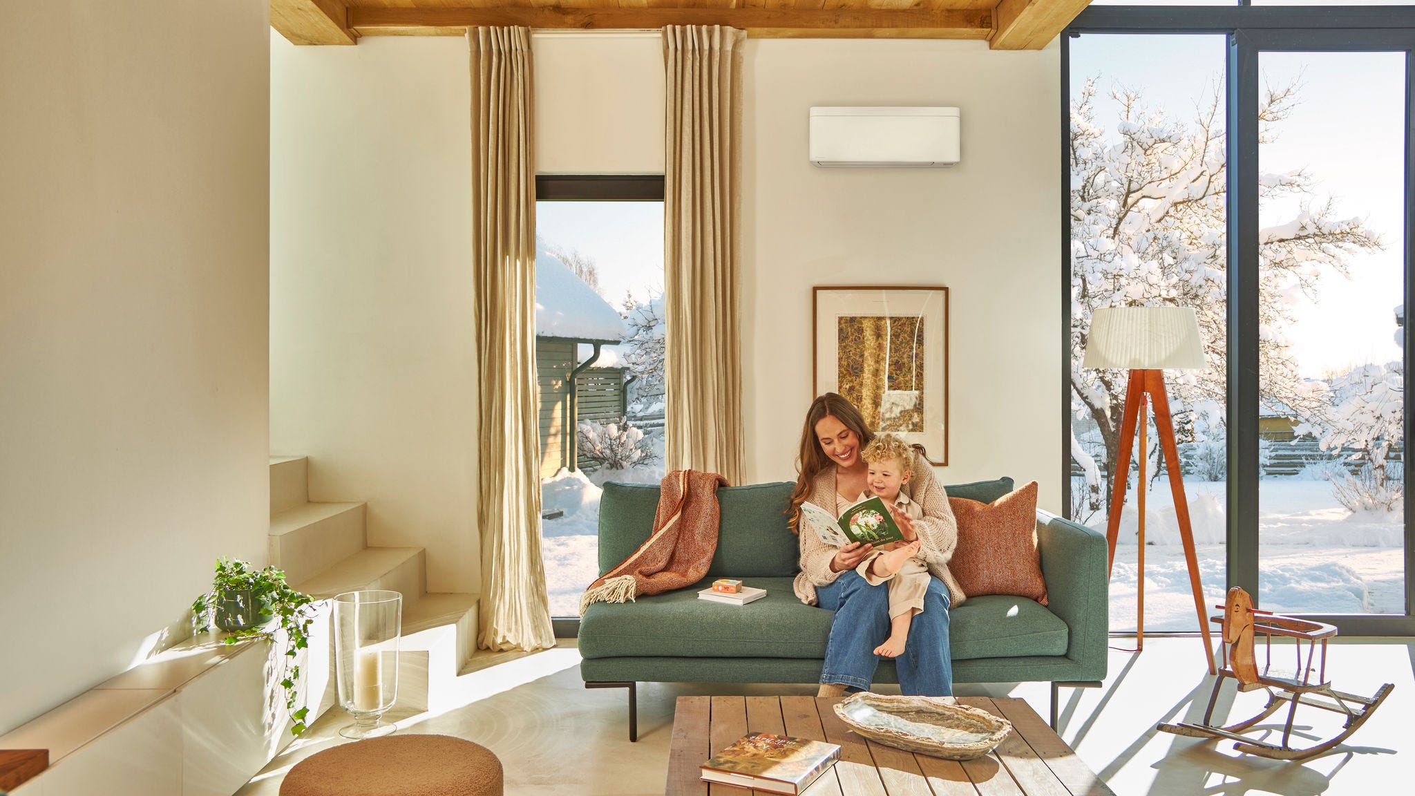 Moeder zit op de bank onder een lucht-luchtwarmtepomp aan de wand en leest voor uit een boek voor haar kind dat op haar schoot zit. Ze genieten samen van de warmte terwijl het buiten sneeuwt.